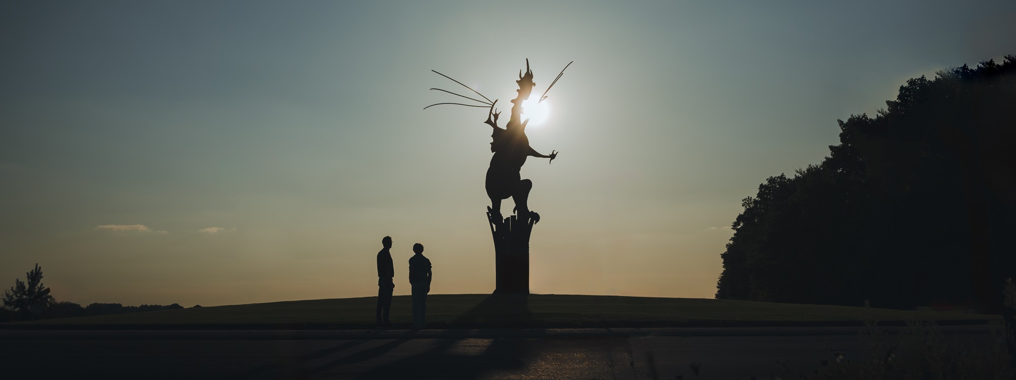Twee mensen bij de draak op de rotonde in Beesel, gefotografeerd bij zonsondergang als onderdeel van de campagne De Verhalen van Hier