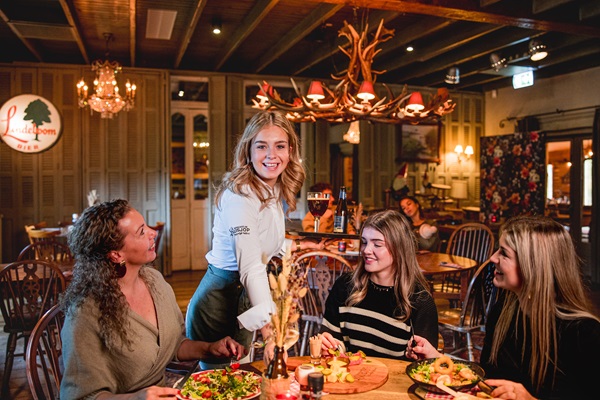 Eine Kellnerin bringt einer Gruppe von drei lächelnden Frauen im Boscafé de Busjop in Heyhuysen mit Holzinterieur und Kronleuchtern das Essen.