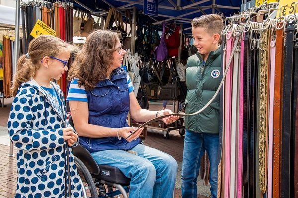 Three people look at and discuss a belt at a market stall at the weekly market in Weert. A woman in a wheelchair holds the belt, while a boy and a girl look on and laugh. Market stalls with bags and accessories can be seen in the background.