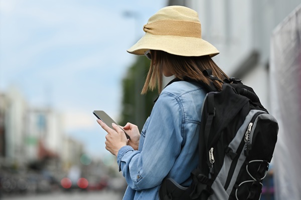 Woman with backpack and hat looks at her smartphone while walking through the city.