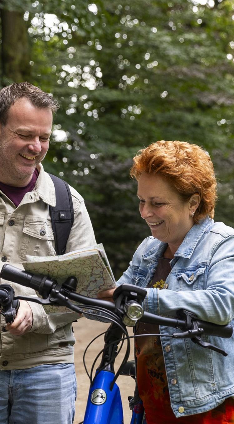 Twee mensen lachen samen terwijl ze met de fiets stilstaan in een bosrijk gebied, kijkend op een fietskaart, met op de achtergrond nog een fietser en kleurrijke bloemen op de voorgrond.