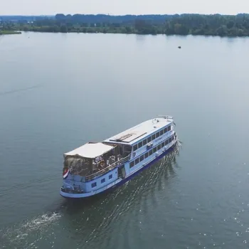Rondvaartboot vaart over de Maas met passagiers aan boord op een zonnige dag, omringd door water en groene oever.