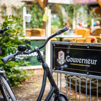 Fiets geparkeerd bij de fietsenstalling met daarop reclame voor Gouverneur bier van Lindeboom met uitzicht op groene tuin en zonnige zitplaatsen op de achtergrond.