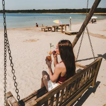 Vrouw geniet van een drankje op een houten schommelbank aan een zandstrand met uitzicht over het Blauwe Meertje in Weert.